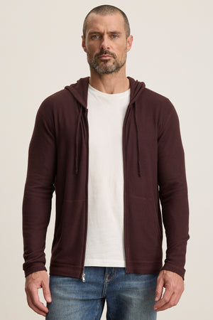 A man stands against a plain background, wearing a white t-shirt, blue jeans, and the LOUDON HOODIE by Velvet by Graham & Spencer—a relaxed fit maroon full-zip hoodie crafted from triblend cozy jersey.
