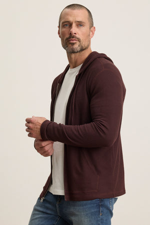 A man with a short beard wears a white t-shirt, blue jeans, and the LOUDON HOODIE by Velvet by Graham & Spencer—a relaxed fit burgundy full-zip hoodie in triblend cozy jersey—as he stands against a plain light background.