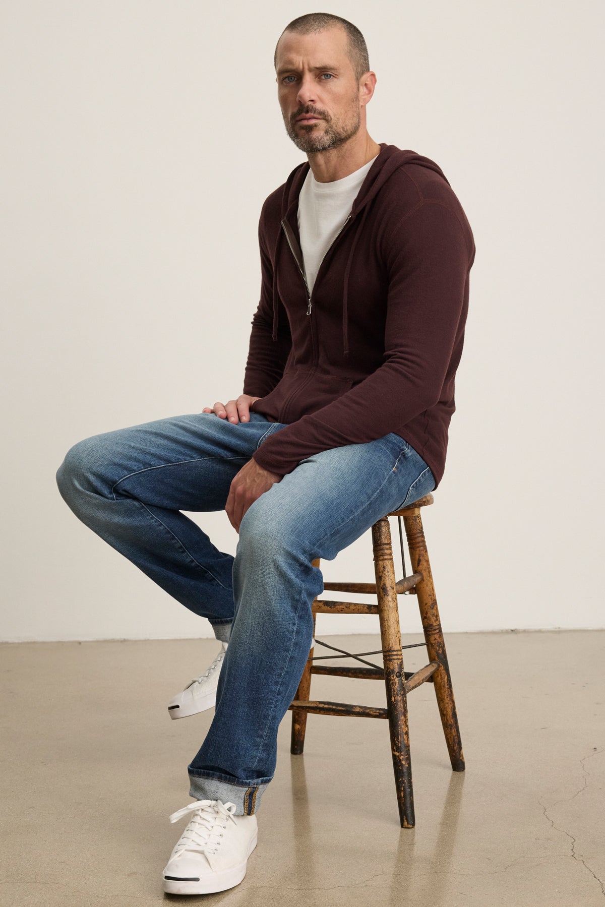 A man with short hair and a beard sits on a wooden stool against a plain background, wearing the LOUDON HOODIE by Velvet by Graham & Spencer—a maroon triblend cozy jersey full-zip hoodie—over a white tee, blue relaxed-fit jeans, and white sneakers.-39905796751553