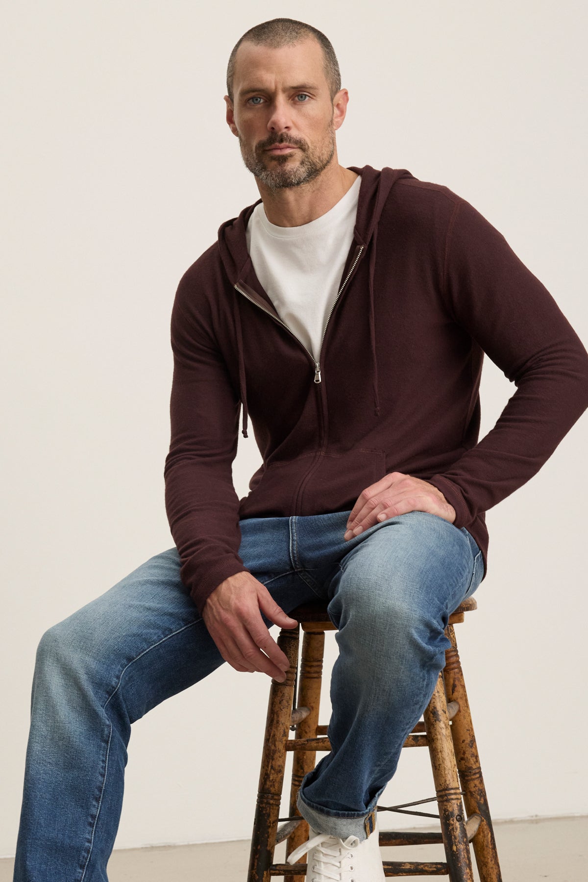 A man with short hair and a beard sits on a wooden stool, wearing the Velvet by Graham & Spencer LOUDON HOODIE in maroon triblend cozy jersey, paired with a white t-shirt, blue jeans, and white sneakers against a plain background.