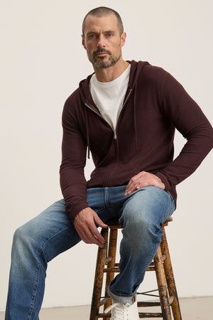 A man with short hair and a beard sits on a wooden stool, wearing the Velvet by Graham & Spencer LOUDON HOODIE in maroon triblend cozy jersey, paired with a white t-shirt, blue jeans, and white sneakers against a plain background.