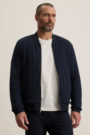A man wearing the Velvet by Graham & Spencer MILES JACKET in navy over a white t-shirt and navy pants stands against a plain light background.