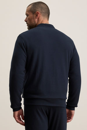 A man with short hair and a beard is seen from behind, wearing the MILES JACKET by Velvet by Graham & Spencer in dark navy with matching pants, set against a plain light background.