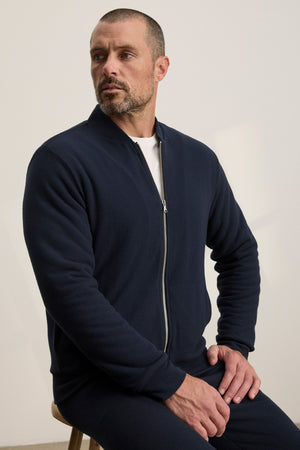 A man with short hair and a beard sits on a stool, wearing the Velvet by Graham & Spencer MILES JACKET in navy over a white shirt, looking to the side.