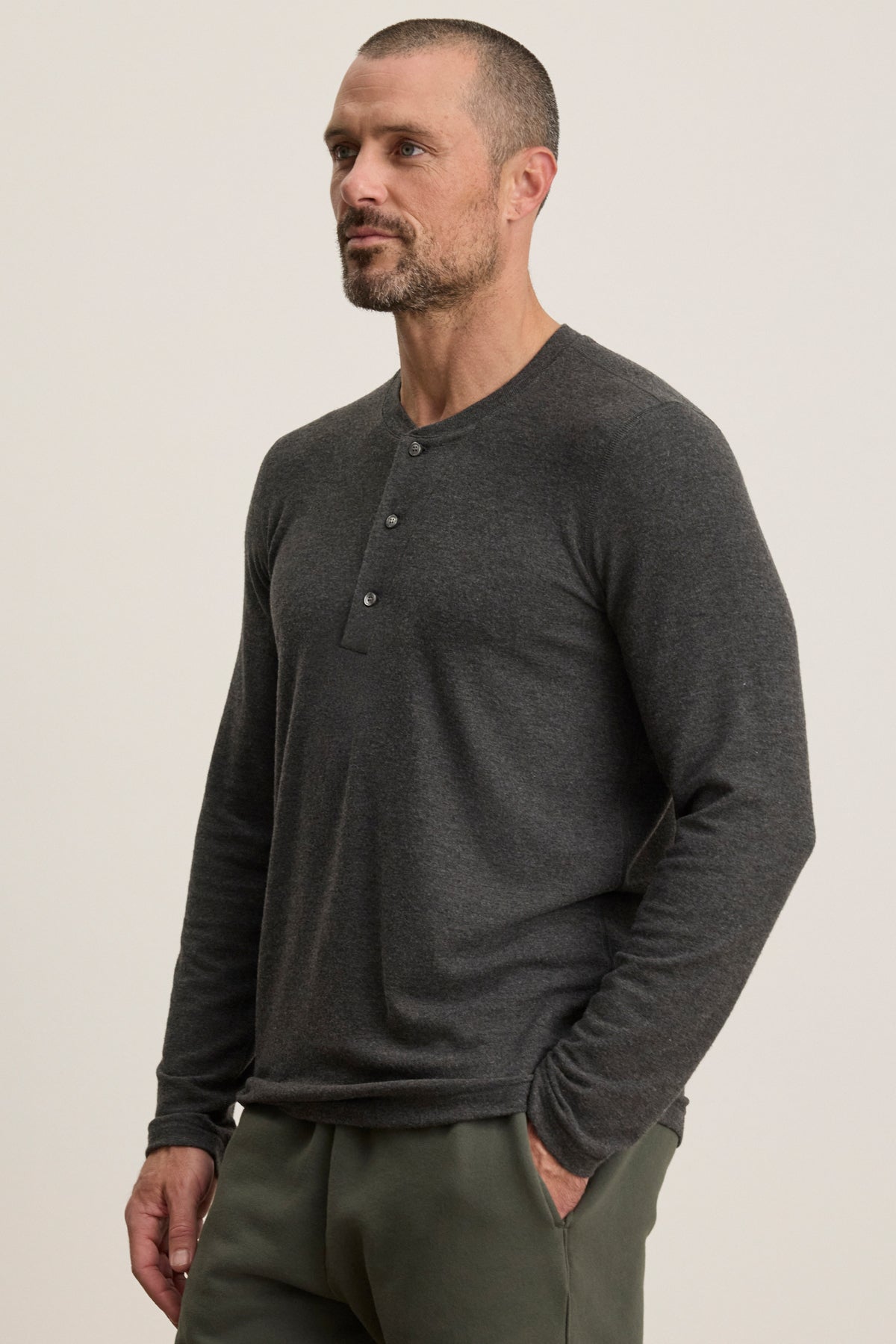 A man with short hair and a beard wears the Velvet by Graham & Spencer RYLAND HENLEY in dark gray jersey with green pants, standing with one hand in his pocket against a plain background—ideal for casual occasions.-40054249062593