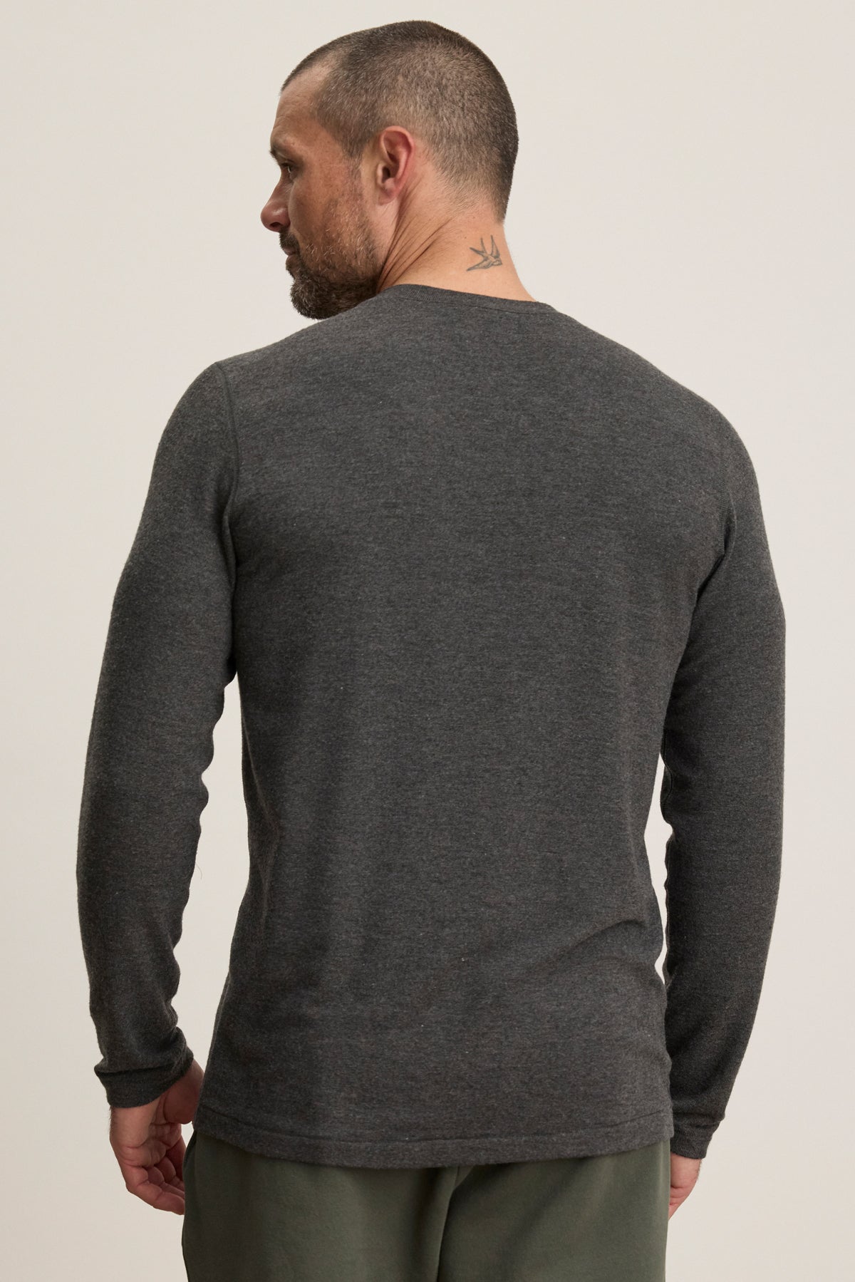 A man with short hair and a neck tattoo wears Velvet by Graham & Spencer’s RYLAND HENLEY, a cozy long-sleeve dark gray 3-button jersey, and green pants, shown from the back—ideal for casual occasions.