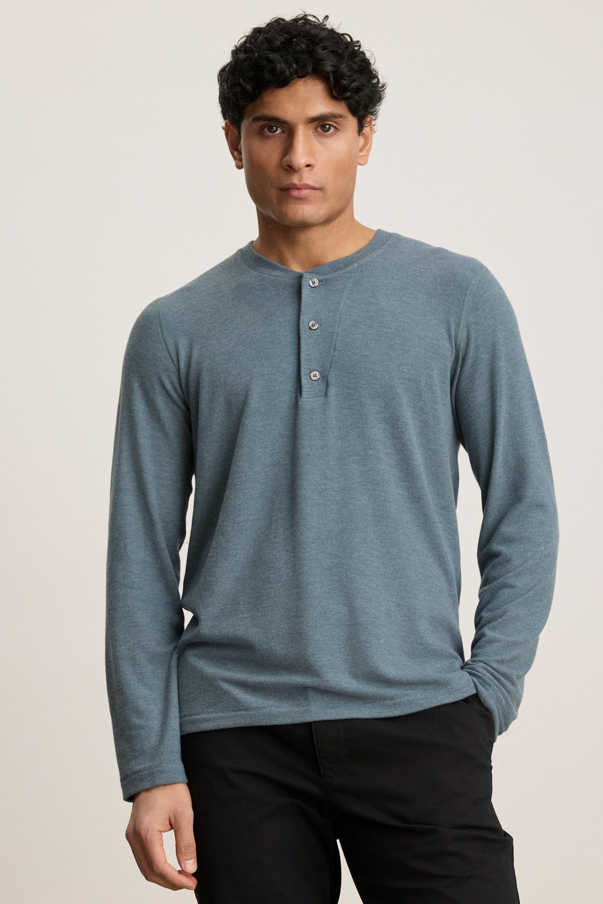 A man with short curly hair wears the RYLAND HENLEY by Velvet by Graham & Spencer in light blue with black pants, standing against a plain light background.