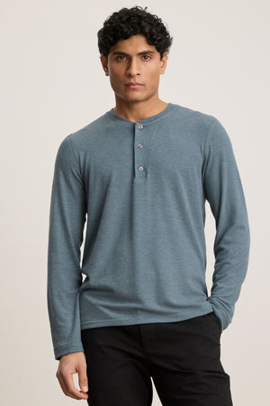 A man with short curly hair wears the RYLAND HENLEY by Velvet by Graham & Spencer in light blue with black pants, standing against a plain light background.