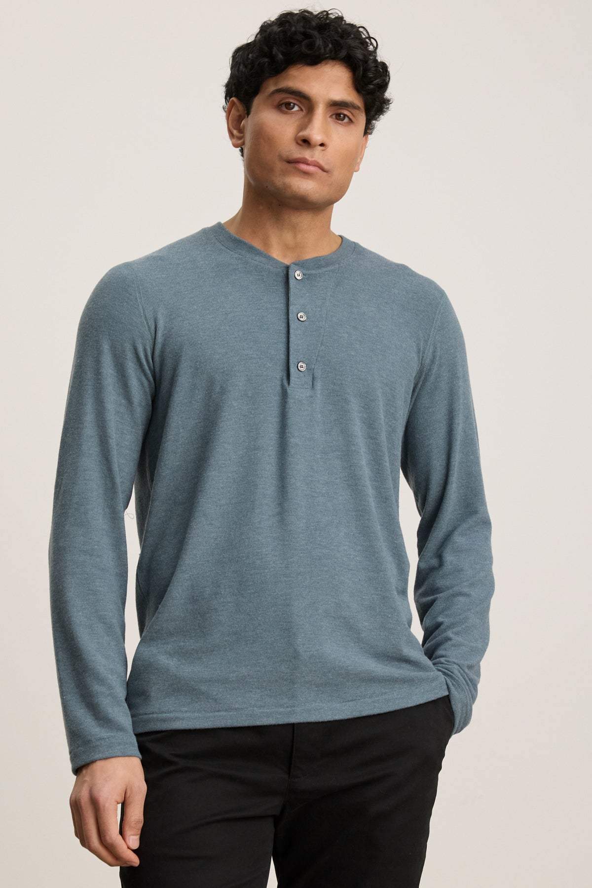 A person wears the Velvet by Graham & Spencer RYLAND HENLEY, a cozy blue long-sleeve jersey shirt, paired with black pants against a plain light background—an ideal casual look.