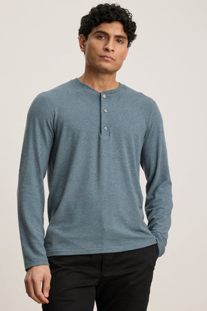 A person wears the Velvet by Graham & Spencer RYLAND HENLEY, a cozy blue long-sleeve jersey shirt, paired with black pants against a plain light background—an ideal casual look.