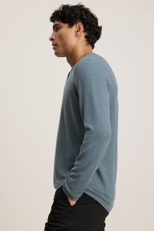 A man with short, curly hair stands in profile against a plain background, wearing the RYLAND HENLEY by Velvet by Graham & Spencer in blue and black pants—ideal for casual occasions.