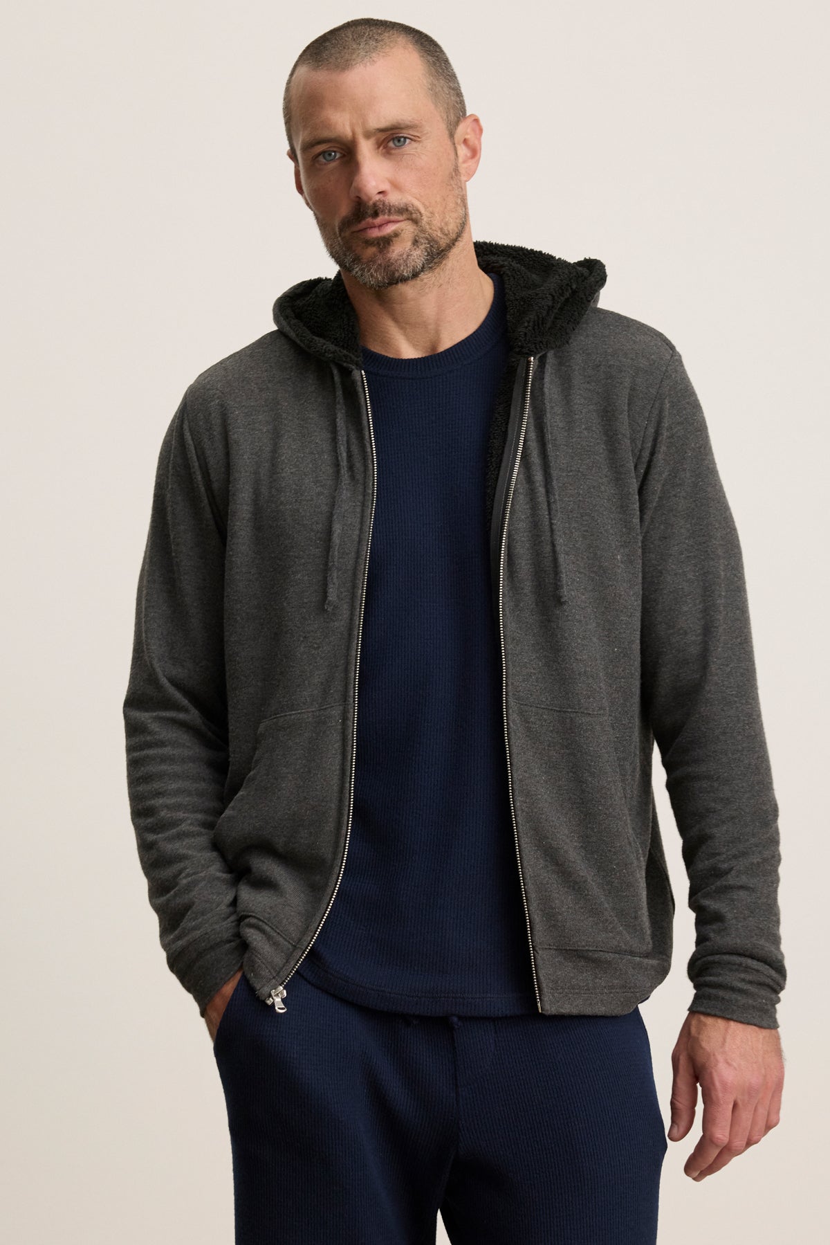 A man with short hair and a beard wears the SALVADORE HOODIE by Velvet by Graham & Spencer, a dark gray zip-up, over a navy shirt and pants, standing against a plain light background.
