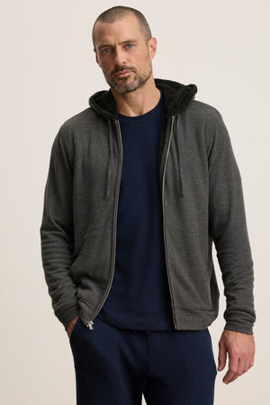 A man with short hair and a beard wears the SALVADORE HOODIE by Velvet by Graham & Spencer, a dark gray zip-up, over a navy shirt and pants, standing against a plain light background.