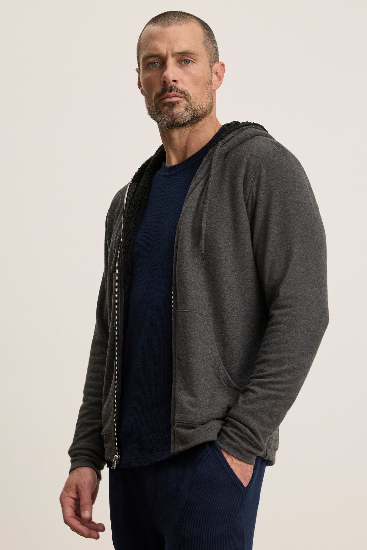 A man with short hair and a beard wears the Velvet by Graham & Spencer SALVADORE HOODIE, a dark grey zip-up style with faux shearling lining, over a navy shirt against a neutral background.