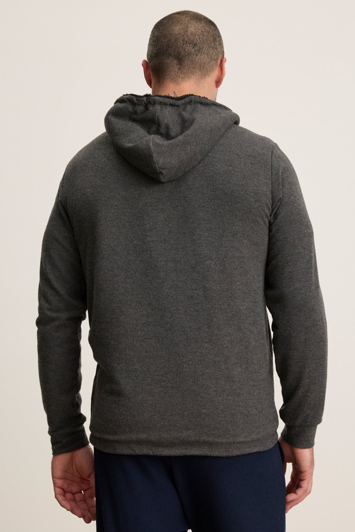 A man is shown from the back against a plain light background, wearing the SALVADORE HOODIE by Velvet by Graham & Spencer—a dark gray hooded sweatshirt with an adjustable drawstring—paired with navy blue pants.
