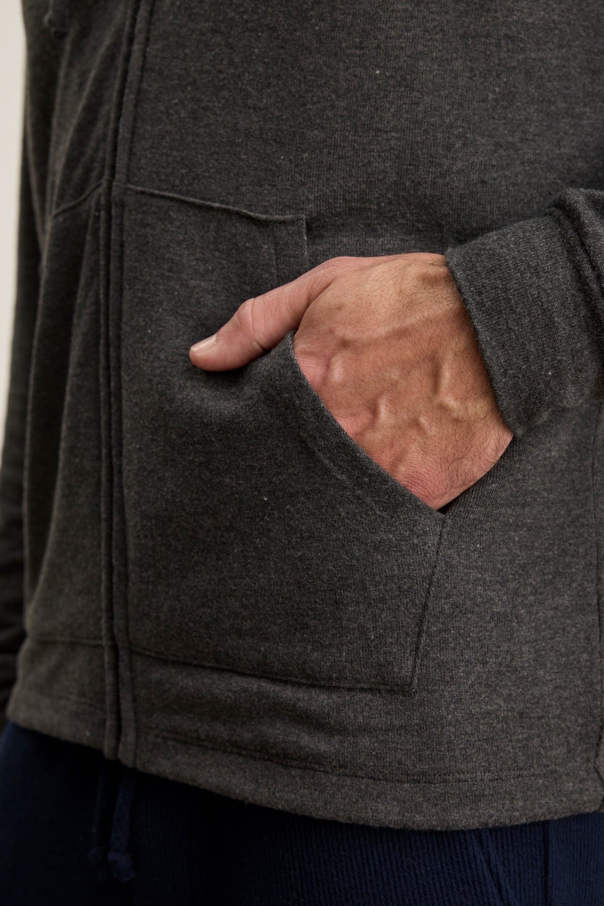 A person’s right hand is partly inside the front pocket of the dark gray SALVADORE HOODIE by Velvet by Graham & Spencer, featuring a zip-up front and an adjustable drawstring hood.
