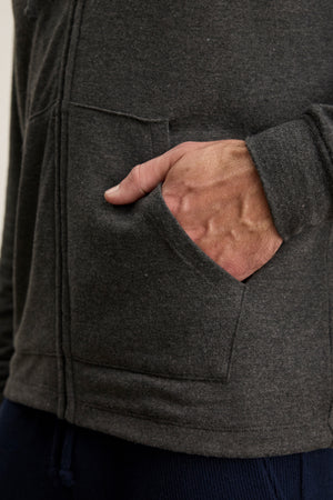 A person’s right hand is partly inside the front pocket of the dark gray SALVADORE HOODIE by Velvet by Graham & Spencer, featuring a zip-up front and an adjustable drawstring hood.