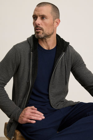 A man with short hair and a trimmed beard sits on a wooden stool against a plain background, wearing the Velvet by Graham & Spencer SALVADORE HOODIE—a dark zip-up style with an adjustable drawstring hood—over a navy shirt.