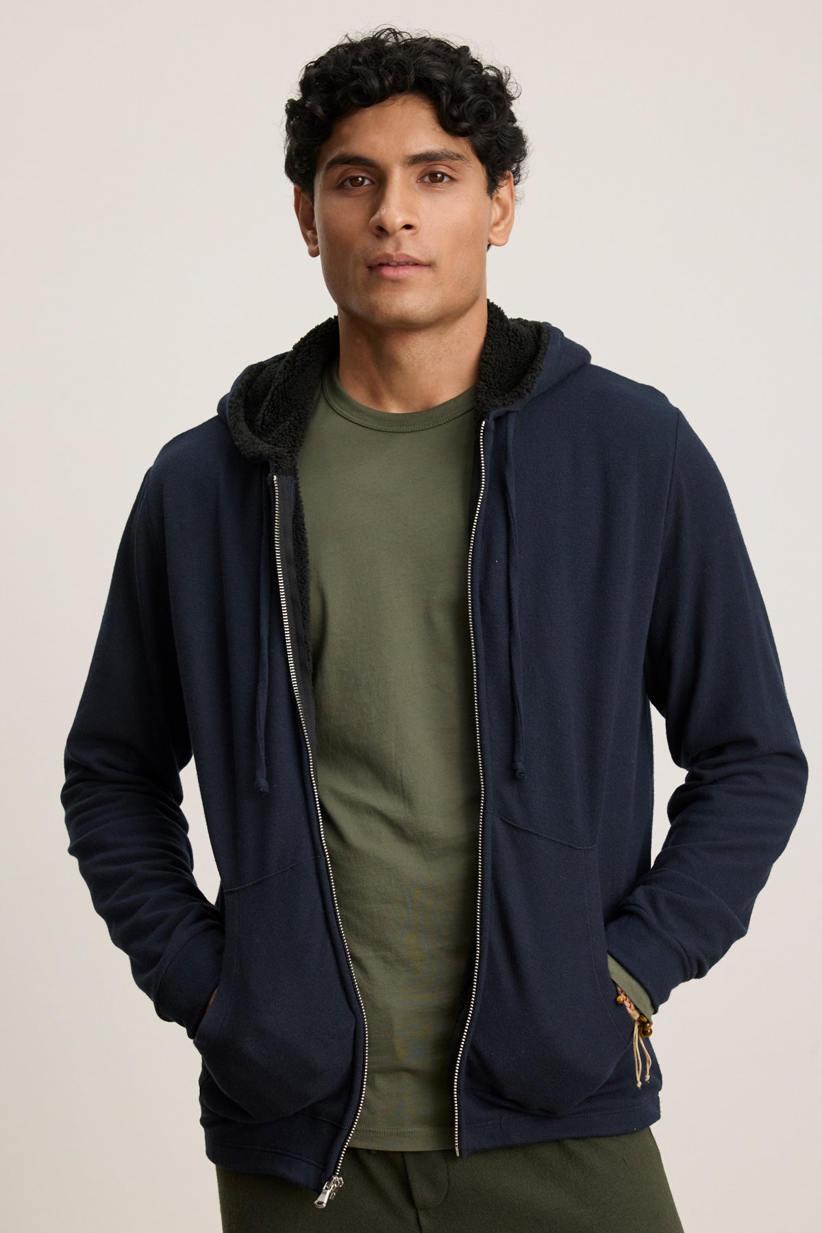 A person faces forward against a neutral background, wearing the Velvet by Graham & Spencer SALVADORE HOODIE in dark blue over an olive green t-shirt, with hands tucked in the hoodie pockets.