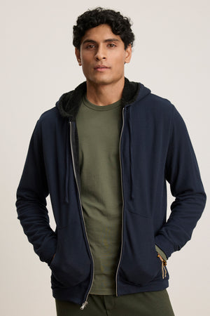 A person faces forward against a neutral background, wearing the Velvet by Graham & Spencer SALVADORE HOODIE in dark blue over an olive green t-shirt, with hands tucked in the hoodie pockets.