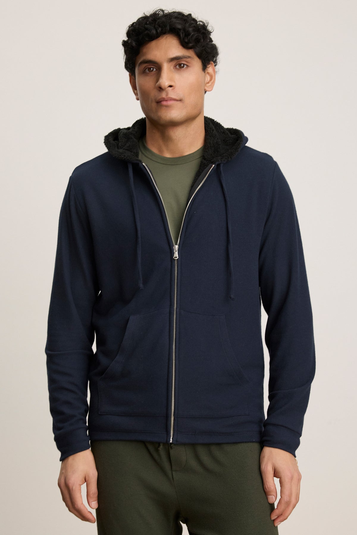 A person wears the SALVADORE HOODIE by Velvet by Graham & Spencer—a navy zip-front hoodie with faux shearling lining—along with a green shirt and pants, standing against a plain background.