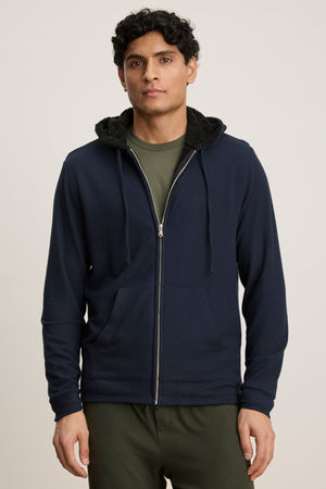A person wears the SALVADORE HOODIE by Velvet by Graham & Spencer—a navy zip-front hoodie with faux shearling lining—along with a green shirt and pants, standing against a plain background.