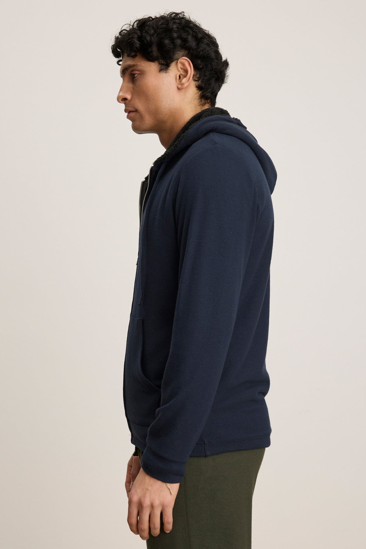A man stands in profile wearing the SALVADORE HOODIE by Velvet by Graham & Spencer—navy blue, zip-up with an adjustable drawstring hood—and olive green pants, set against a plain light background.