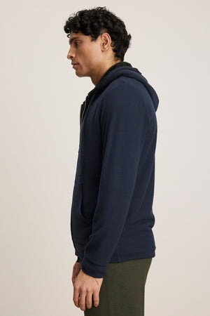 A man stands in profile wearing the SALVADORE HOODIE by Velvet by Graham & Spencer—navy blue, zip-up with an adjustable drawstring hood—and olive green pants, set against a plain light background.