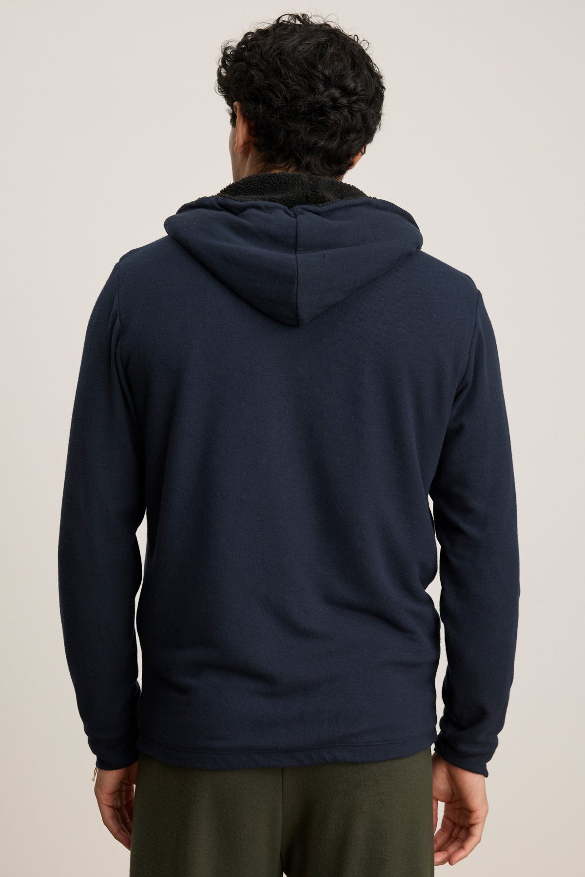 A person with dark, curly hair is seen from the back wearing the Velvet by Graham & Spencer SALVADORE HOODIE in navy blue with an adjustable drawstring hood, paired with dark green pants against a plain light background.