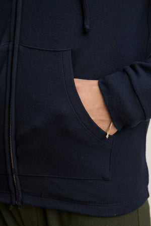 A person wearing the SALVADORE HOODIE by Velvet by Graham & Spencer in navy blue, featuring a zip-up front, adjustable drawstring hood, and their right hand in the front pocket.
