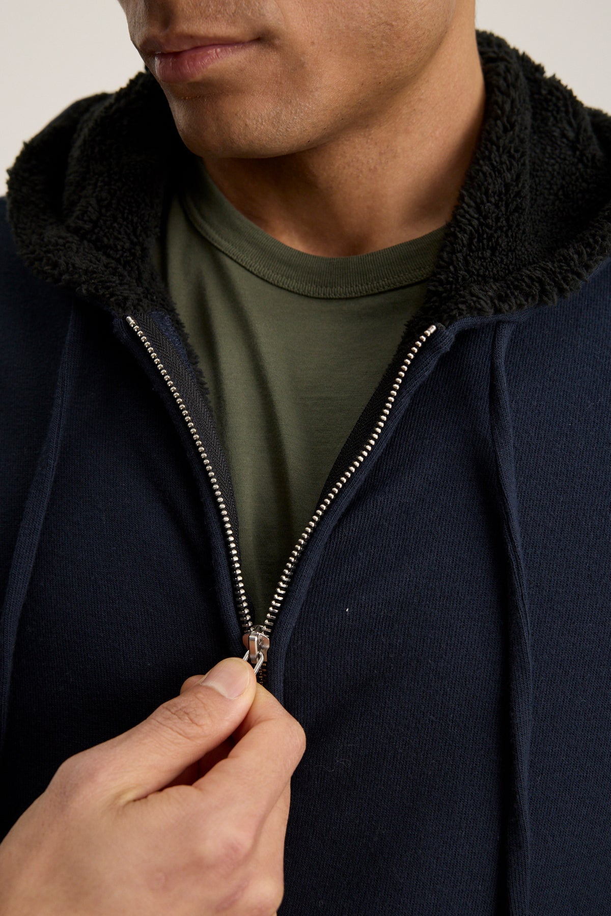 A person zips up the SALVADORE HOODIE by Velvet by Graham & Spencer, featuring a navy blue color, adjustable drawstring hood, black fleece lining, and an olive green t-shirt underneath.