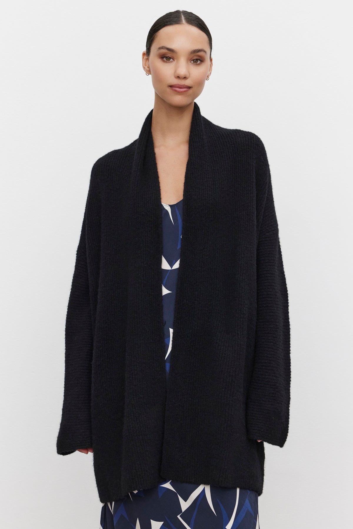 A person wearing a CALLI CARDIGAN by Velvet by Graham & Spencer over a patterned dress stands against a plain white background.