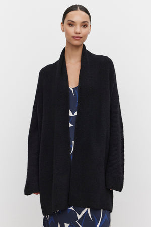 A person wearing a CALLI CARDIGAN by Velvet by Graham & Spencer over a patterned dress stands against a plain white background.