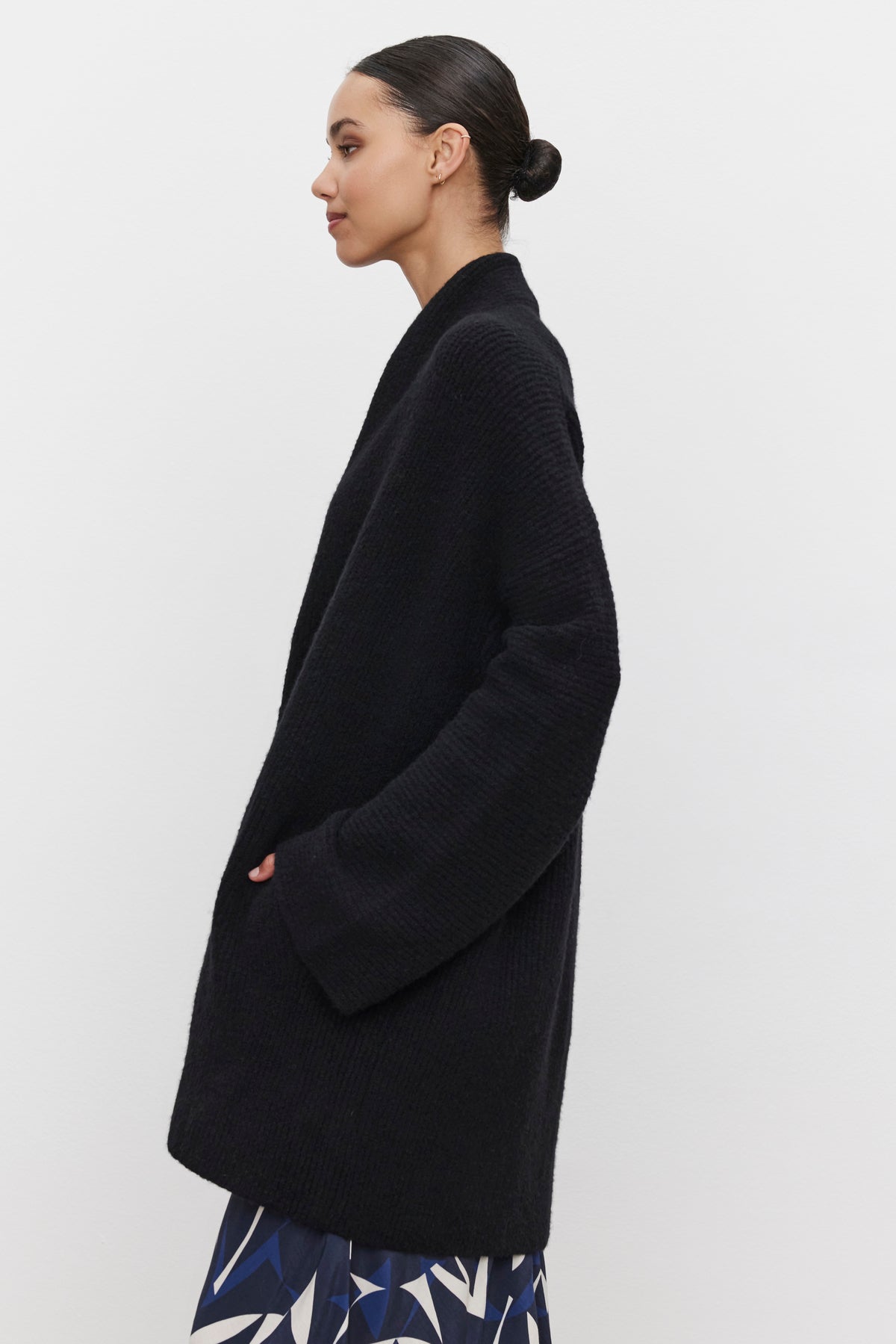 A woman with dark hair in a bun, wearing an oversized black coat over a printed skirt, stands against a white background, her ensemble completed by the elegant CALLI CARDIGAN from Velvet by Graham & Spencer.-37676875874497