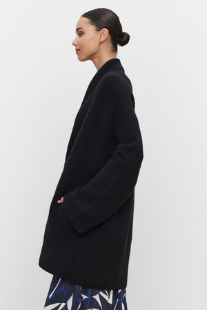 A woman with dark hair in a bun, wearing an oversized black coat over a printed skirt, stands against a white background, her ensemble completed by the elegant CALLI CARDIGAN from Velvet by Graham & Spencer.