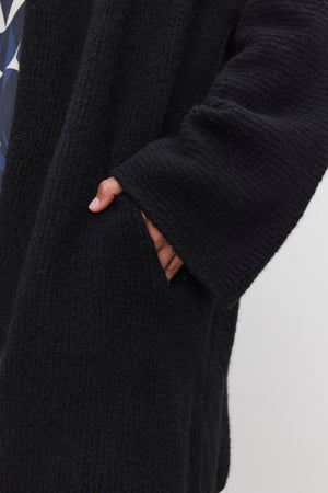 A person wearing the CALLI CARDIGAN by Velvet by Graham & Spencer, with its luxurious black texture and hand casually placed inside the pocket, evoking the timeless elegance of a shawl collar cardigan.