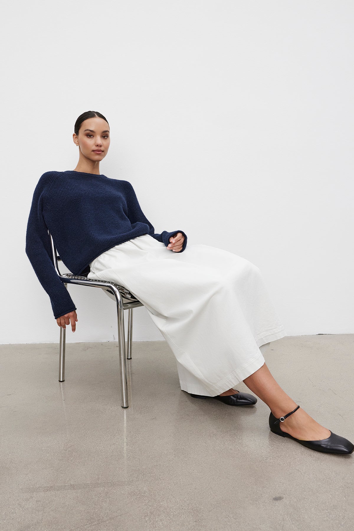 A person sitting on a chair against a white background, wearing a navy blue sweater, the SOPHIA SKIRT by Velvet by Graham & Spencer, and black flat shoes.