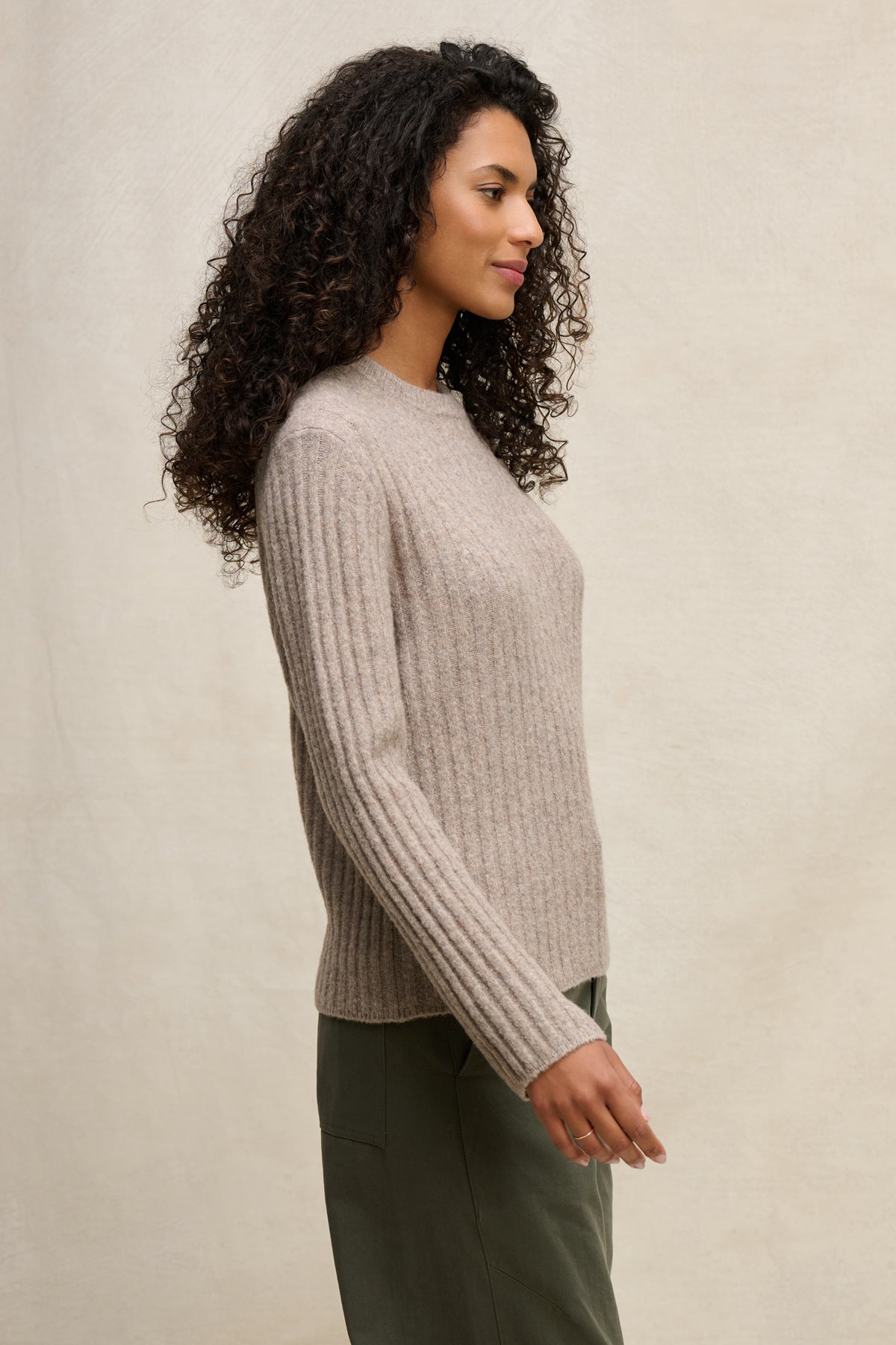 A woman with curly hair stands in profile, wearing the IMANA SWEATER by Velvet by Graham & Spencer—a cozy wool blend in reverse jersey stitch—and olive green pants against a neutral background.-39838351392961
