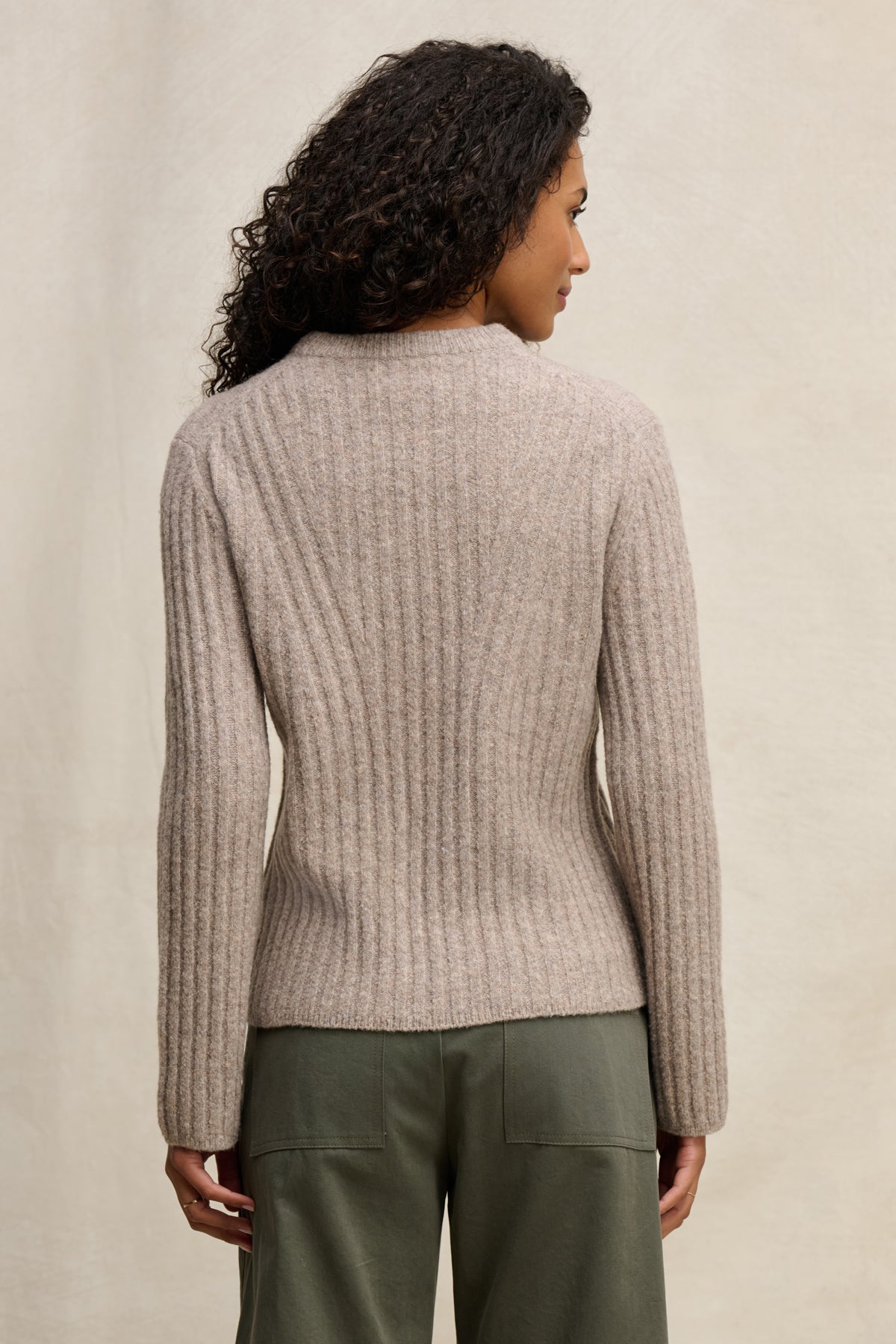 A person with curly hair is shown from the back, wearing the IMANA SWEATER by Velvet by Graham & Spencer—a cozy ribbed beige wool blend—paired with olive green pants against a neutral background.-39838351360193