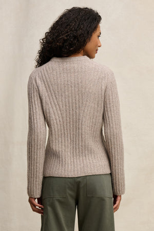 A person with curly hair is shown from the back, wearing the IMANA SWEATER by Velvet by Graham & Spencer—a cozy ribbed beige wool blend—paired with olive green pants against a neutral background.