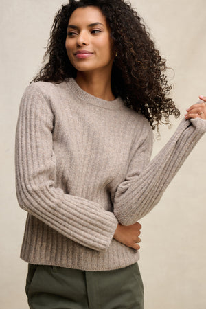A woman with curly hair wears the IMANA SWEATER by Velvet by Graham & Spencer in a cozy wool blend, paired with olive green pants, standing against a neutral background and gazing slightly to the side.