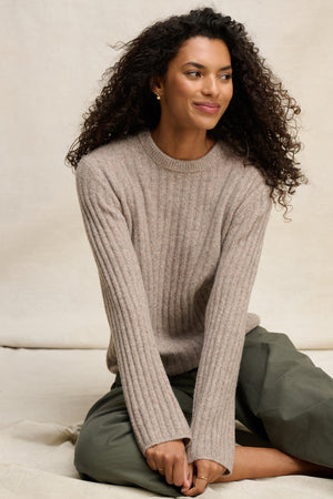 A woman with long curly hair wears the IMANA SWEATER by Velvet by Graham & Spencer, featuring ribbed details, styled with green pants as she sits on the floor against a neutral background.