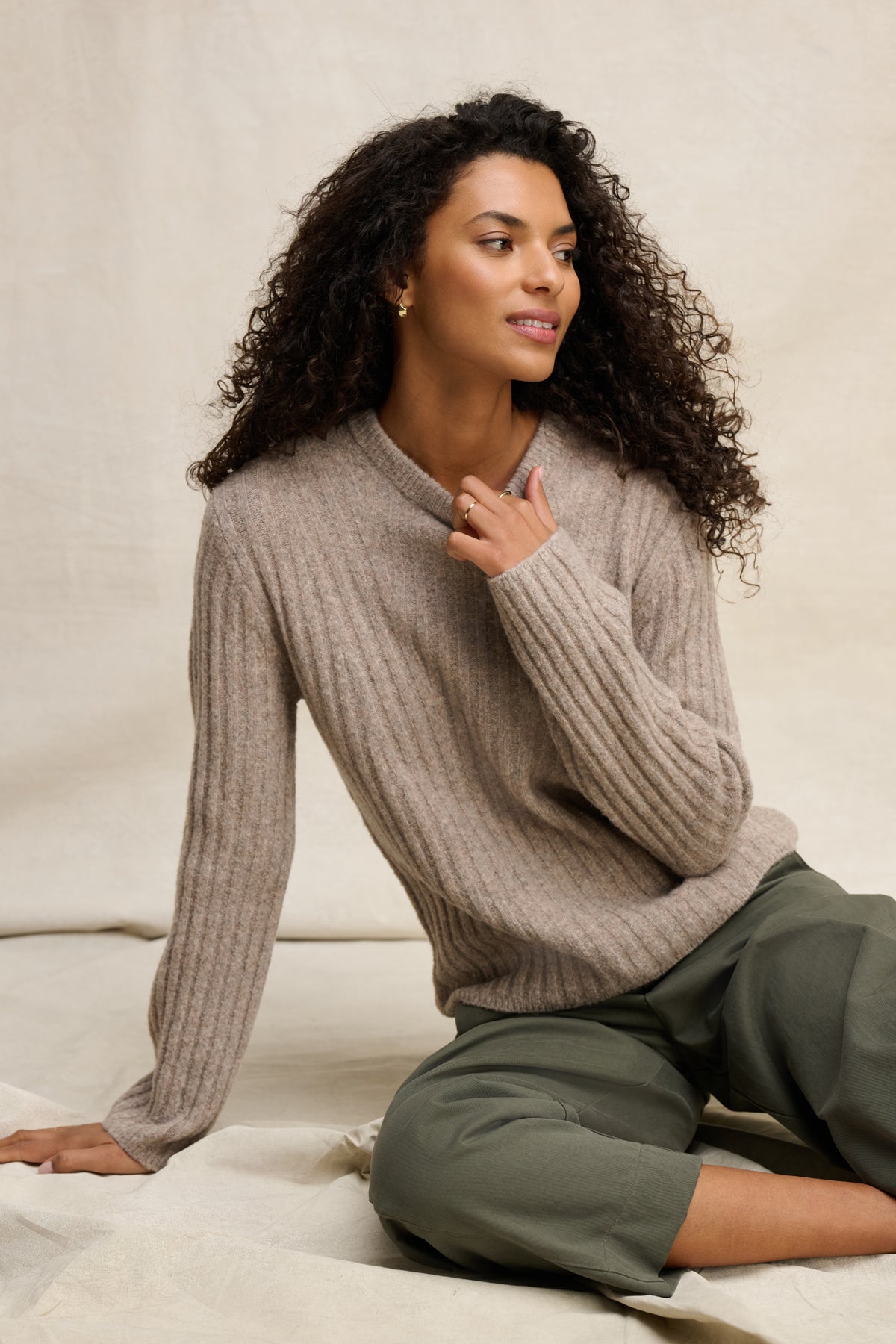A woman with curly hair wears the IMANA SWEATER by Velvet by Graham & Spencer— a cozy beige wool blend in ribbed reverse jersey stitch—paired with olive green pants, seated on a light fabric background and gazing slightly to the side.