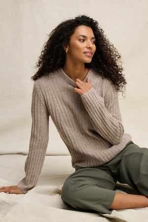 A woman with curly hair wears the IMANA SWEATER by Velvet by Graham & Spencer— a cozy beige wool blend in ribbed reverse jersey stitch—paired with olive green pants, seated on a light fabric background and gazing slightly to the side.