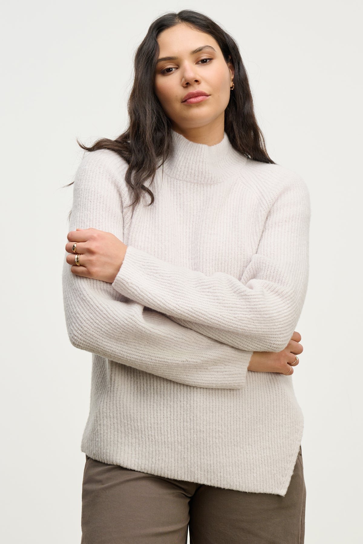 A woman with long dark hair stands against a plain background, wearing the light-colored LESA SWEATER by Velvet by Graham & Spencer and brown pants, arms crossed and expression neutral.-39838357717185