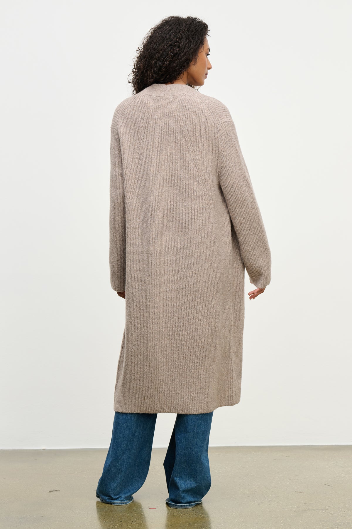 A person with curly hair stands with their back to the camera, wearing the MISHA CARDIGAN by Velvet by Graham & Spencer and blue jeans against a plain light background.-40046297481409