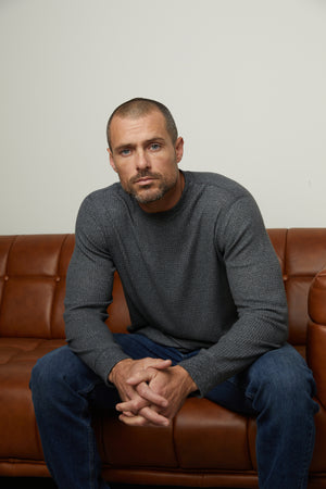 A man sitting on a brown leather couch, perfect for cooler days, wearing the Velvet by Graham & Spencer PONCHO THERMAL CREW.