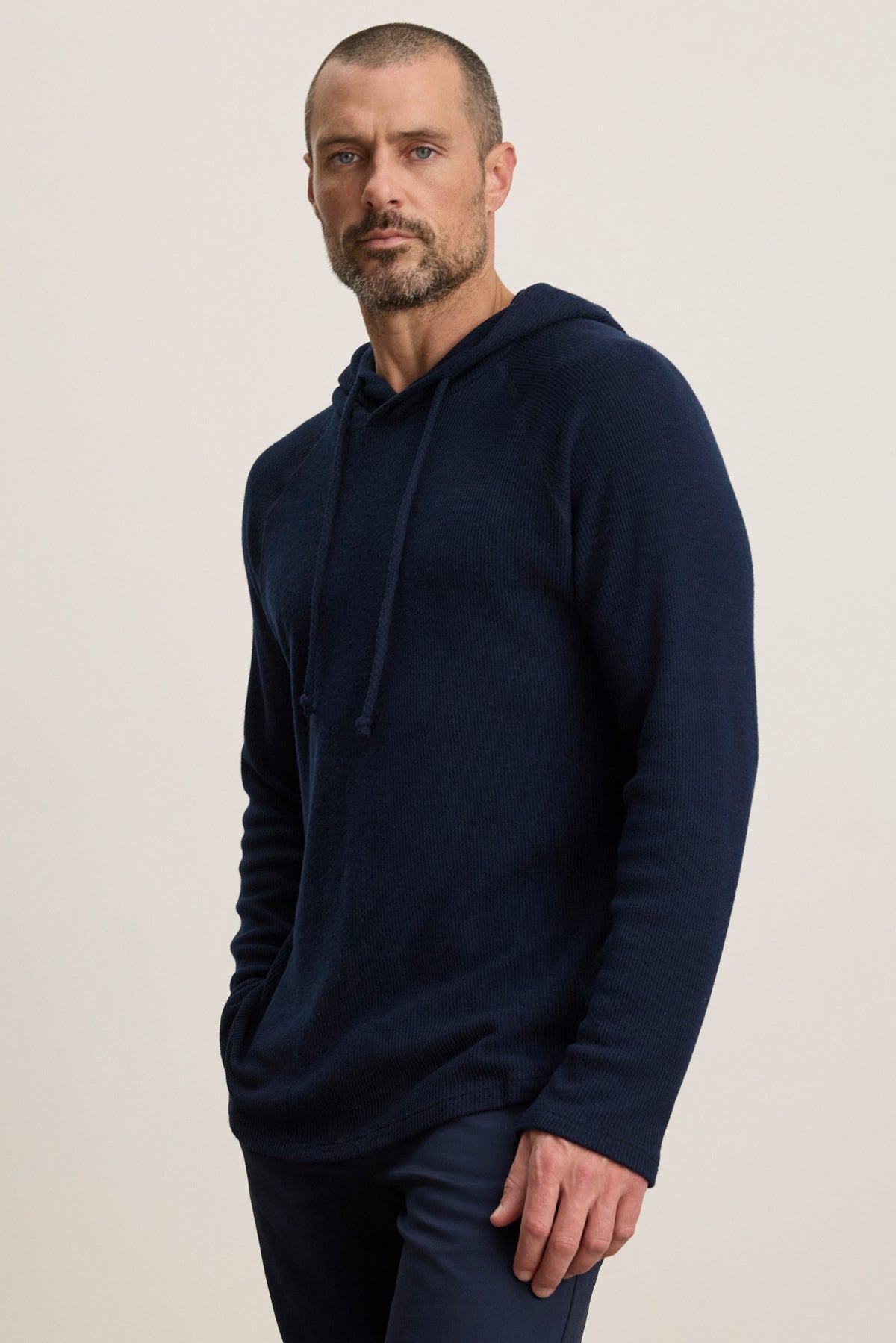 A man with short hair and a beard wears the Velvet by Graham & Spencer ROMEO HOODIE—a relaxed fit, navy blue pullover—paired with dark pants, standing against a plain light background.-39957110653121