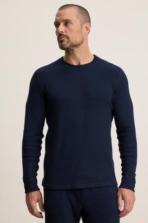 A man with short hair and a trimmed beard wears the TOBIAS CREW by Velvet by Graham & Spencer— a cozy, long-sleeve dark blue textured thermal shirt with matching pants, standing against a plain light background.