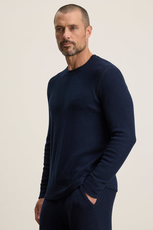 Man with short hair and trimmed beard wears the TOBIAS CREW by Velvet by Graham & Spencer— a fitted, long-sleeve navy blue thermal sweater— with matching pants, standing against a plain light background.